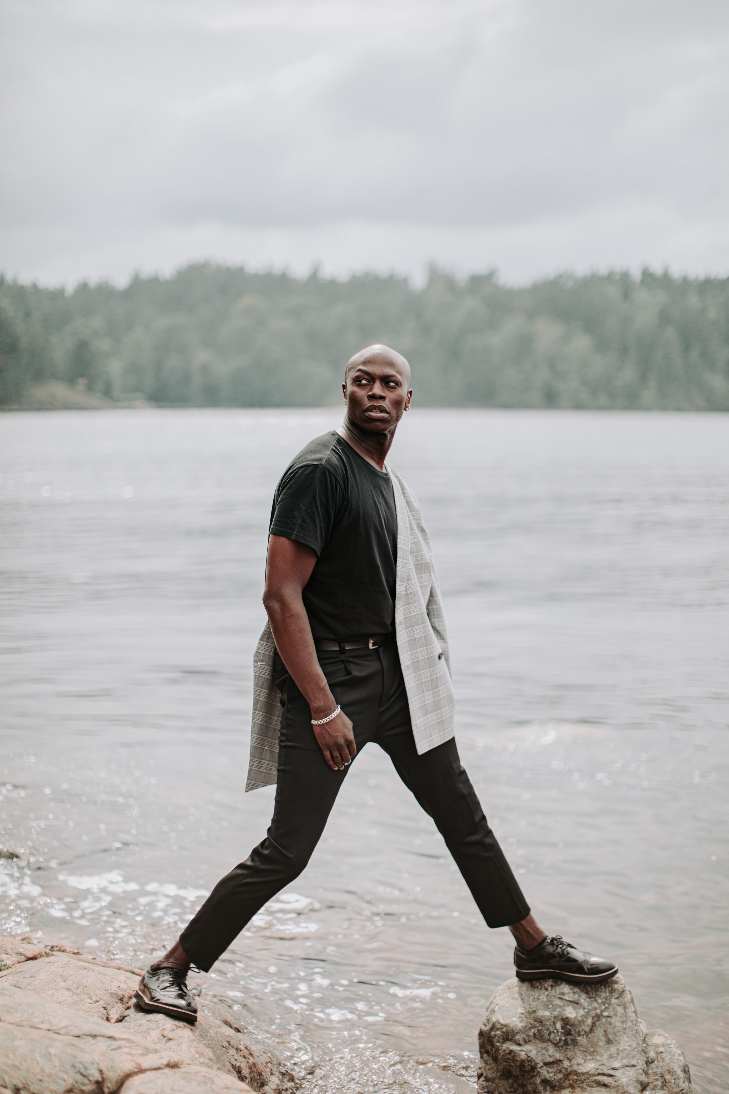 Man posing on rocks by a lake in black tee and draped blazer